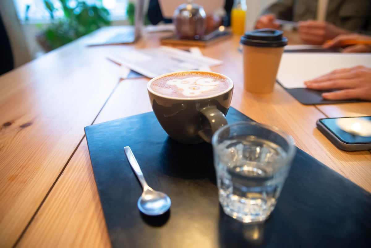A cappuccino and a glass of water on a wooden meeting table, with laptops, papers and a takeaway cup around it