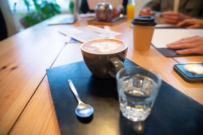 A cappuccino and a glass of water on a wooden meeting table, with laptops, papers and a takeaway cup around it